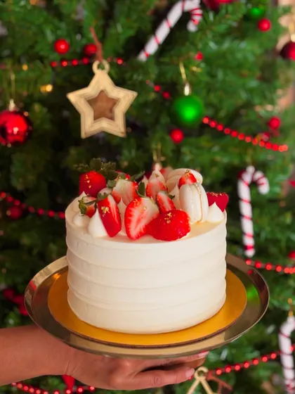 A festive Christmas cake with white buttercream, topped with fresh strawberries and macarons, held in front of a decorated Christmas tree.
