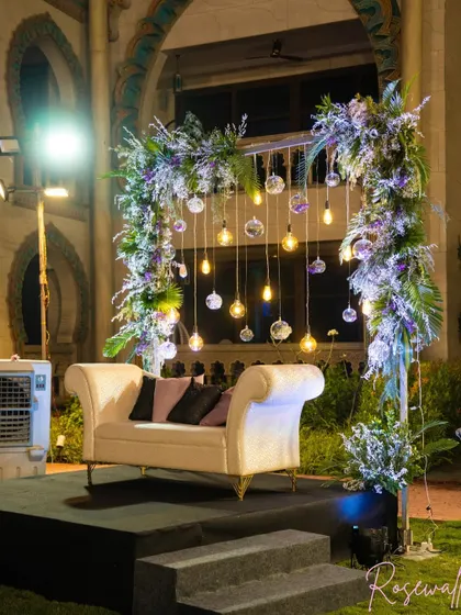 A wider view of the special seating arrangement for the couple of honor. The elegant white loveseat is set on a raised platform, framed by beautiful flowers and lights.