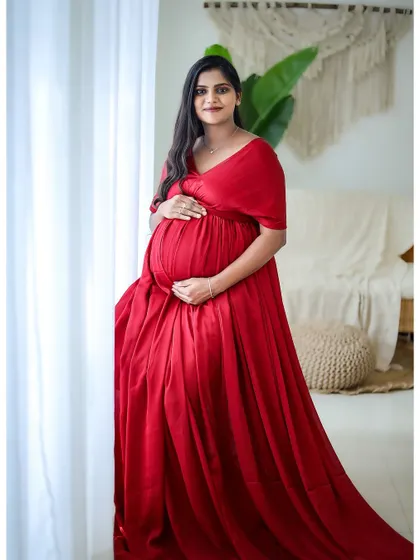 A simple and elegant portrait of a mom-to-be in a classic red gown, standing by a window for soft, natural lighting.