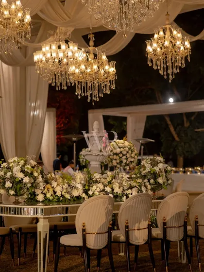 A close-up of the head table at the white-themed reception. The mirrored tabletop is adorned with a runner of white roses and orchids, while the water fountain and chandeliers create a symphony of light, sound, and fragrance.