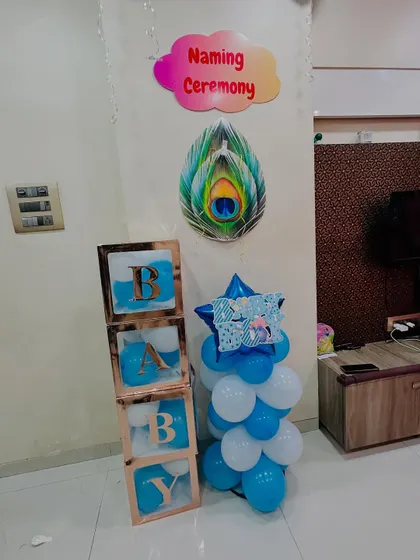 A detailed shot of the decor elements for a baby boy's naming ceremony. It includes custom 'BABY' blocks filled with blue balloons and a wall decorated with a 'Naming Ceremony' sign and peacock feather art.