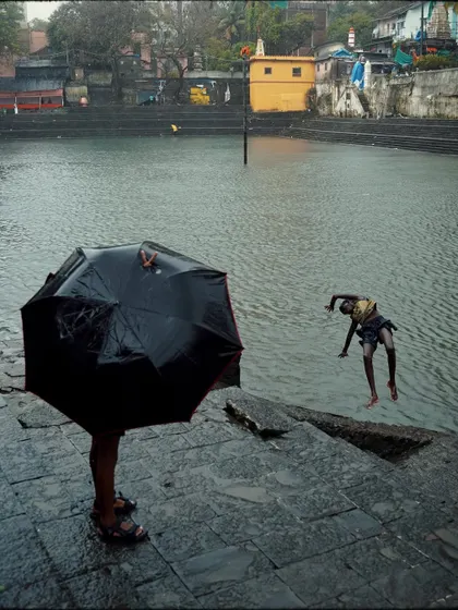 A study in contrasts at Banganga Tank: one person stands shielded by an umbrella while another leaps joyfully into the water.