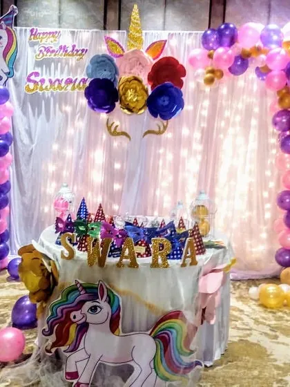A whimsical unicorn birthday party for Swara. This setup features a unicorn head with paper flowers, a light-strung curtain backdrop, and balloon pillars in purple and pink.
