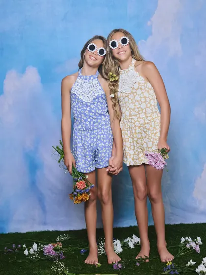 A studio shot for a girls' summer collection. The faux grass, flowers, and cloud backdrop create a whimsical, outdoor feel in a controlled environment.
