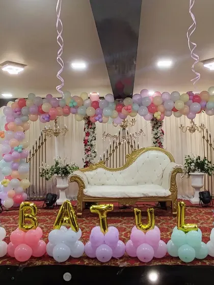 An elegant and grand first birthday setup in a party hall. This design includes a wide pastel-colored organic balloon arch over a regal white chaise lounge, with the name "BATUL" spelled out in balloon letters at the front.