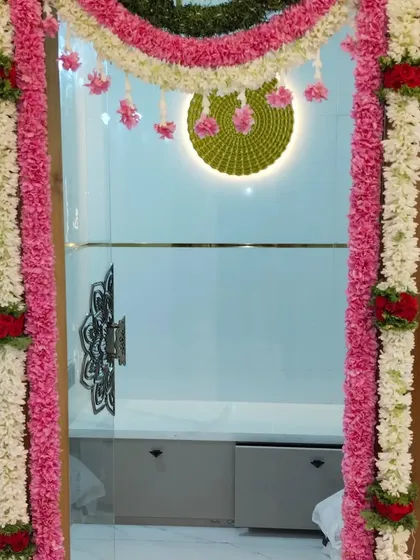 I often blend traditional decor with modern homes. Here, a beautiful floral frame made of pink, white, and red flower strings decorates the main glass door for a housewarming pooja, adding a touch of elegance and tradition.