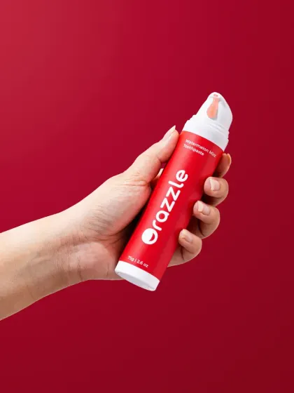 A hand-held shot of Orazzle's watermelon toothpaste, demonstrating the product's pump feature. This is a utility-focused shot that clearly communicates how the product works.