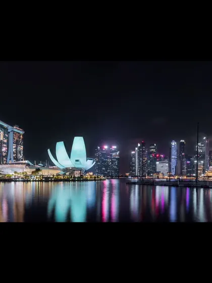 A panoramic night photograph of Singapore's Marina Bay, capturing the iconic skyline and its reflection in the water.
