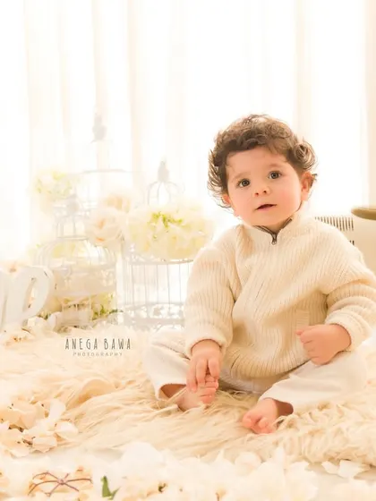 A sweet and simple moment from a first birthday session. The baby sits on a soft fur rug, surrounded by flowers, looking directly at the camera.