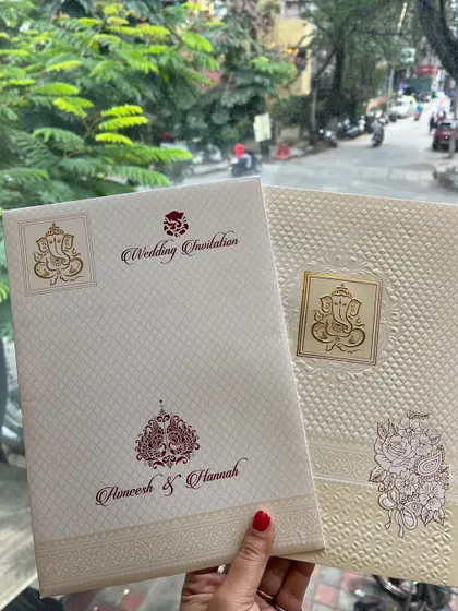 A pair of elegant, off-white invitations featuring a Ganesha motif. The cards use high-quality textured paper and subtle gold foil for a premium feel.