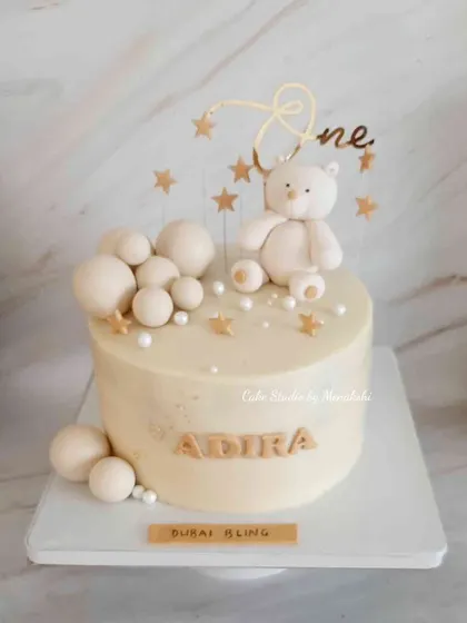 A monochrome first birthday cake for Adira. This elegant all-white design is decorated with a teddy bear, stars, and bubbles for a chic celebration.