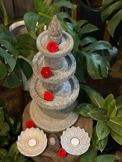 A granite temple lamp decorated with red flowers, ready for a festival. These tiered stone lamps are a classic element of Indian decor, perfect for adding a touch of tradition to your garden.