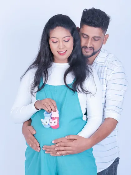 A joyful couple portrait from a studio maternity session. They hold up tiny baby booties, one pink and one blue, in a cute and playful pose.