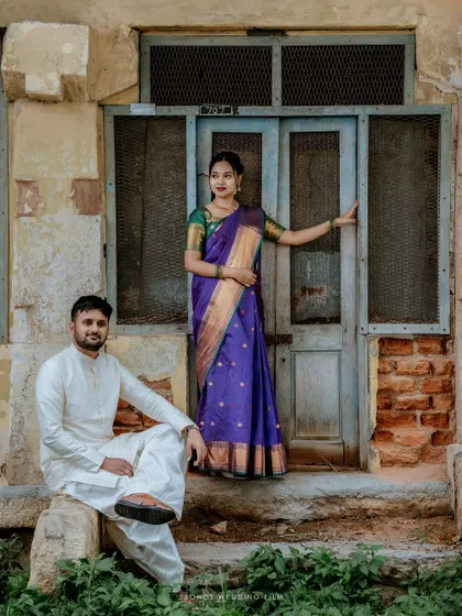 A striking portrait of the couple in traditional outfits, the bride standing elegantly in a doorway while the groom is seated, creating a classic, regal composition.