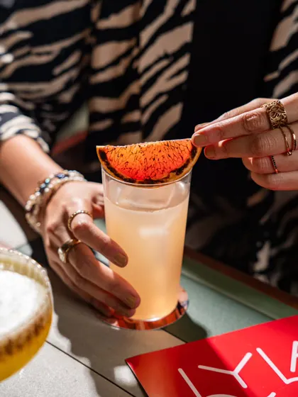 A close-up of a hand garnishing a drink with a dehydrated blood orange slice at Lyla Bombay. This highlights the fresh, craft element of their cocktails.
