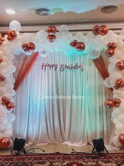 An elegant arch for an 89th birthday, using white and rose gold chrome balloons with a fabric backdrop and professional lighting.