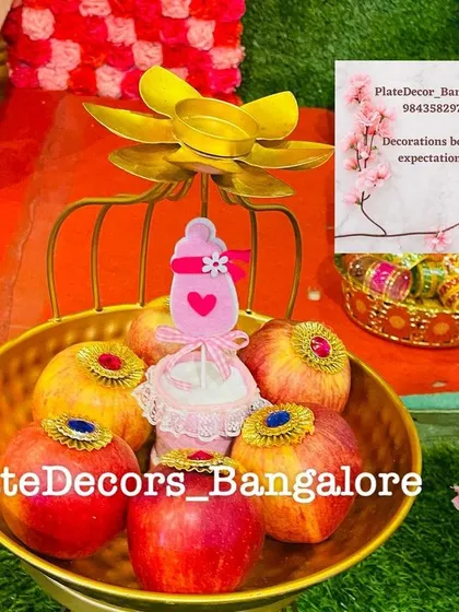 Decorated apples in a golden lotus-style basket with a pink baby bottle cutout. This is a great option for a baby girl's seemantham ceremony.