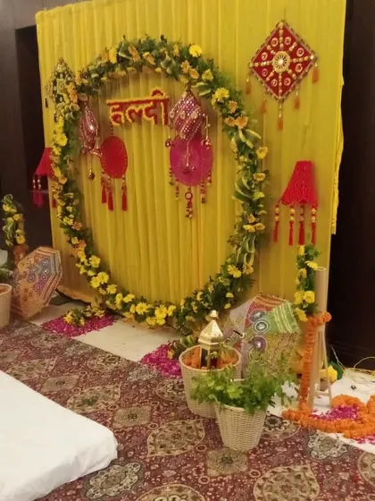 An indoor Haldi setup featuring a circular floral frame against a yellow draped wall. The decor includes traditional props and baskets filled with flowers.