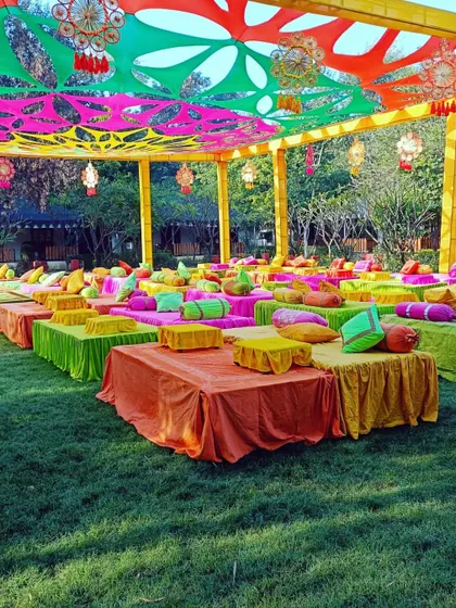 A vibrant outdoor Mehendi setup with low seating and colorful cushions under a canopy of bright orange, pink, and green drapes.