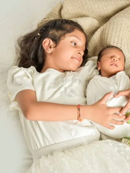 A tender moment between siblings, as an older sister lies down and gently holds her swaddled newborn sibling, looking at them with love and wonder.