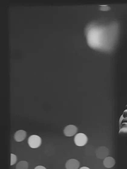 A close-up black and white portrait of a woman's face as she looks up, with soft bokeh lights in the background. The minimalist composition and gentle lighting create a quiet, hopeful mood.
