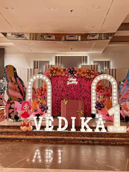 A butterfly-themed stage for Vedika's first birthday, with a pink flower wall, an Eiffel Tower prop, and large marquee letters.