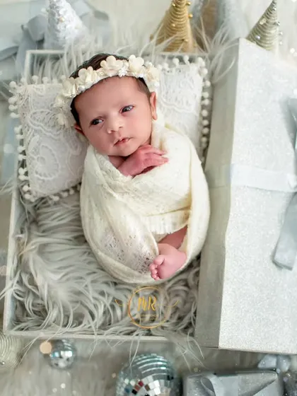 A beautifully wrapped newborn lies in a gift box, surrounded by sparkling silver disco balls and Christmas decorations.