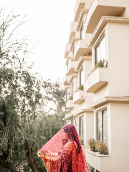 A low-angle shot of the bride in her beautiful red saree, with the building in the background, creating a sense of grandeur.