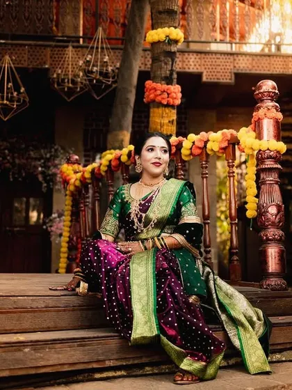 A full-length portrait of a Maharashtrian bride seated on the steps of a traditionally decorated wooden structure. She looks regal and elegant in her rich nauvari saree.