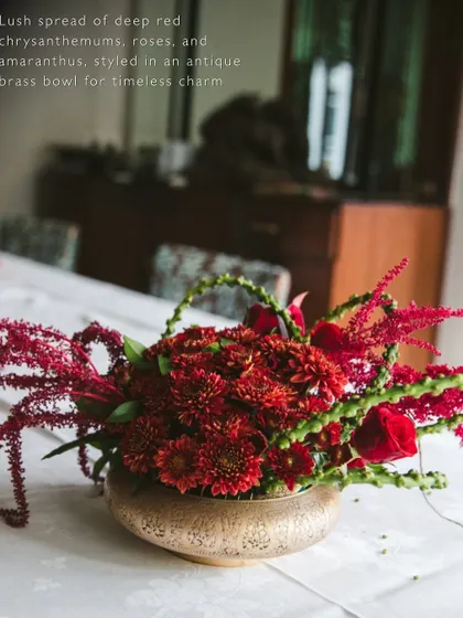 A lush arrangement of deep red chrysanthemums, roses, and amaranthus in an antique brass bowl, adding timeless charm to a tablescape.