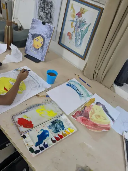 A view of the artist's workspace. You can see the watercolor palette with bright yellows and blues, test swatches, and the painting in progress.