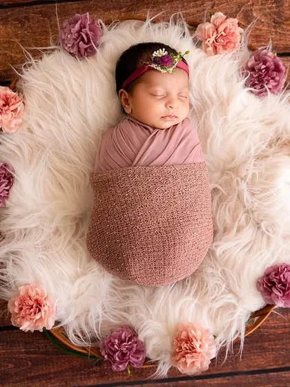 A sleeping newborn, wrapped and wearing a floral headband, is surrounded by a circle of flowers on a soft, white fur rug.