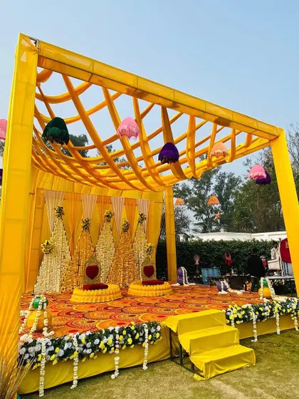 A complete Haldi stage setup in a garden. The yellow canopy has a unique lattice design, and the stage is decorated with marigold backdrops and floral arrangements, ready for the ceremony.