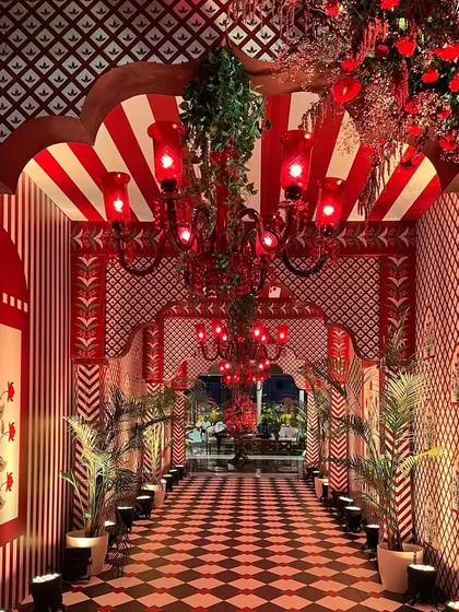A lavish cultural-themed entrance, with a passageway defined by red and white patterned walls, checkered flooring, and ornate red chandeliers, evoking the feeling of walking through a royal Indian palace.