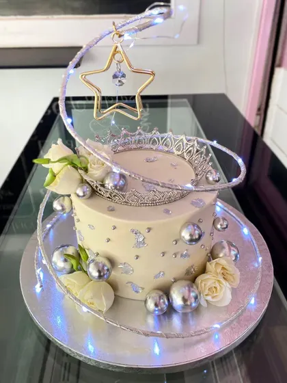 A glamorous cake fit for royalty, featuring a silver crown, silver spheres, and white roses. The design is wrapped in a swirl of fairy lights, adding a magical, sparkling touch.