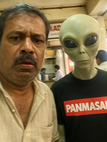 A man takes a disgruntled selfie with an alien wearing a 'PANMASALA' t-shirt. A classic 'Riyal Life' image, finding humor in the mundane collision of the otherworldly and the everyday.