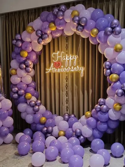 A first-anniversary party with a full circular balloon arch in shades of purple. A "Happy Anniversary" neon sign and a number '1' foil balloon complete the look.