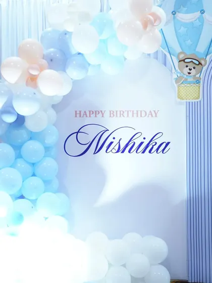A close-up of a teddy bear themed backdrop, personalized with the baby's name and surrounded by a soft blue and white balloon arch.