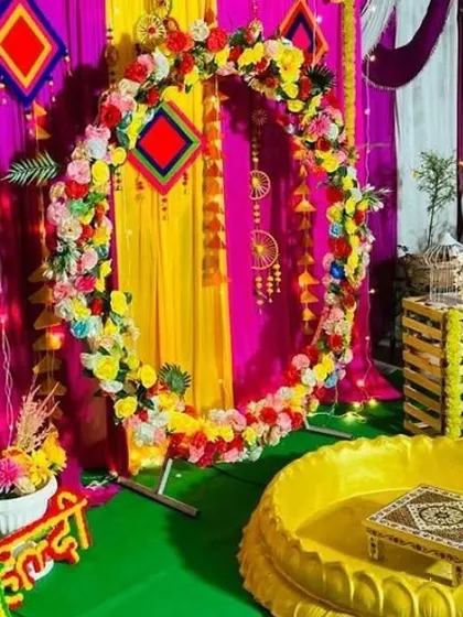 A vibrant Haldi ceremony setup with a circular floral arch against a backdrop of pink and yellow drapes. The decor includes a golden 'urli' and traditional hanging elements.