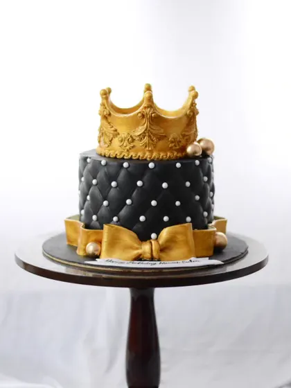 A cake fit for a king. This black and gold cake features a quilted fondant pattern and is topped with a hand-sculpted golden crown and bow.