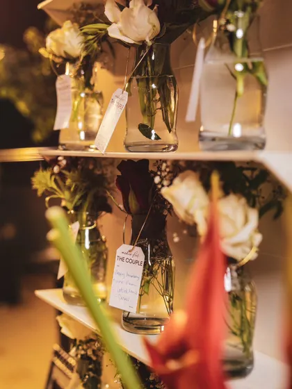 A close-up of the wishing well, showing the small glass bottles with handwritten notes and single stems of white roses. It's a delicate and personal touch for an anniversary celebration.