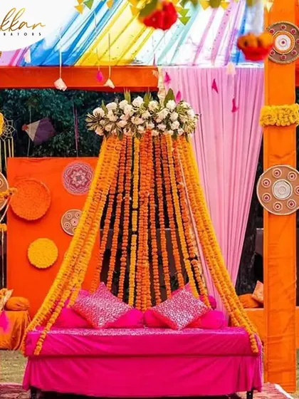 A vibrant Mehendi seating arrangement with a canopy of marigold strings. The bright pink sofa contrasts beautifully with the orange decor, creating a lively and colorful space.