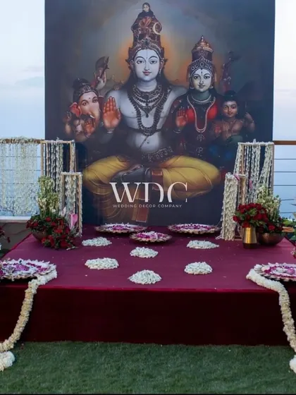 A sacred space for rituals, with a backdrop featuring a beautiful painting of Lord Shiva and his family. The area is adorned with garlands of jasmine and plates of flower petals.