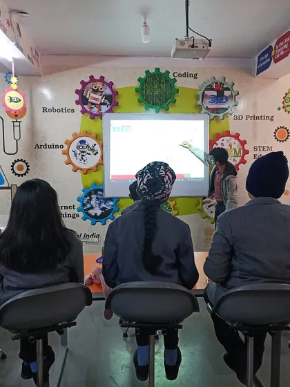 An instructor points to a smartboard in a classroom decorated with themes like Robotics, Coding, and 3D Printing, outlining the day's lesson for the students.