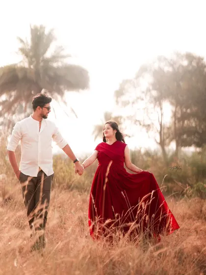 A romantic shot of a couple walking through a field of tall grass. The mother-to-be's red dress and the hazy, warm light create a dreamy, cinematic feel.
