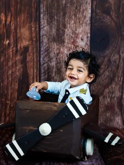 A smiling toddler dressed as a pilot sits in a wooden airplane prop. The rustic wood background complements the vintage aviation theme of this creative kids' portrait session.