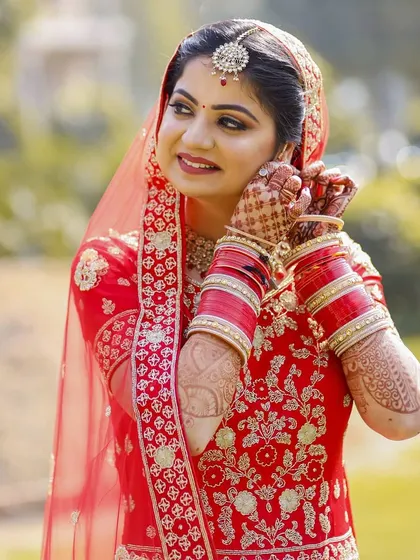 This outdoor bridal portrait captures a quiet, candid moment. The bride is adjusting her earring, a simple gesture that feels natural and elegant. The soft, natural light beautifully illuminates her red embroidered suit and the intricate henna on her hands.