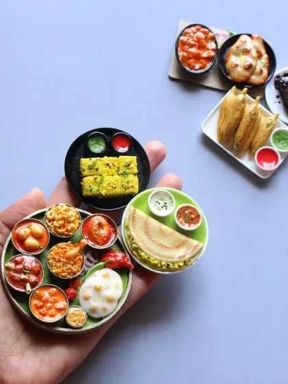 A hand holding a variety of miniatures, including a non-veg thali, dosa, and dhokla.