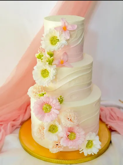 A beautiful three-tier cake with a soft pink ombre wave texture. The design is completed with a delicate arrangement of pink and white gerbera daisies and carnations.