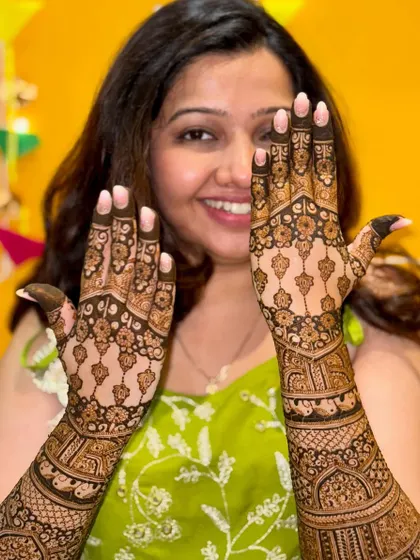 A happy bride showing off her beautiful back-of-hand mehendi. The symmetrical design features delicate hanging motifs and intricate patterns, creating a very elegant look.
